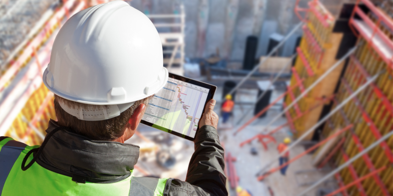 Construction worker in safety gear reviewing building plans on a tablet at a job site.